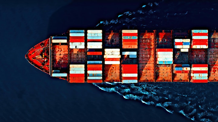 Aerial view of a cargo ship carrying colorful containers, cutting through deep blue waters with visible wave patterns.