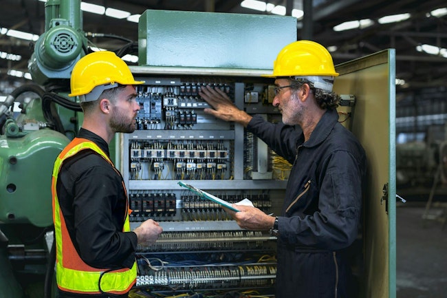 Technician checking operation of control cabinet of production machinery in industrial factory. The technician teaches the new mechanic the job of checking operation of the mechanical control cabinet.