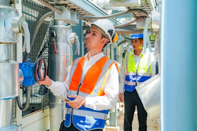 Two workers are examining machinery in an industrial facility. One person, wearing a white shirt and hard hat, operates a control panel, while the other observes in a bright safety vest.