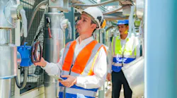 Two workers are examining machinery in an industrial facility. One person, wearing a white shirt and hard hat, operates a control panel, while the other observes in a bright safety vest.