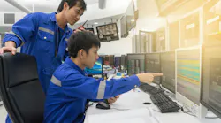 Two workers are examining machinery in an industrial facility. One person, wearing a white shirt and hard hat, operates a control panel, while the other observes in a bright safety vest.