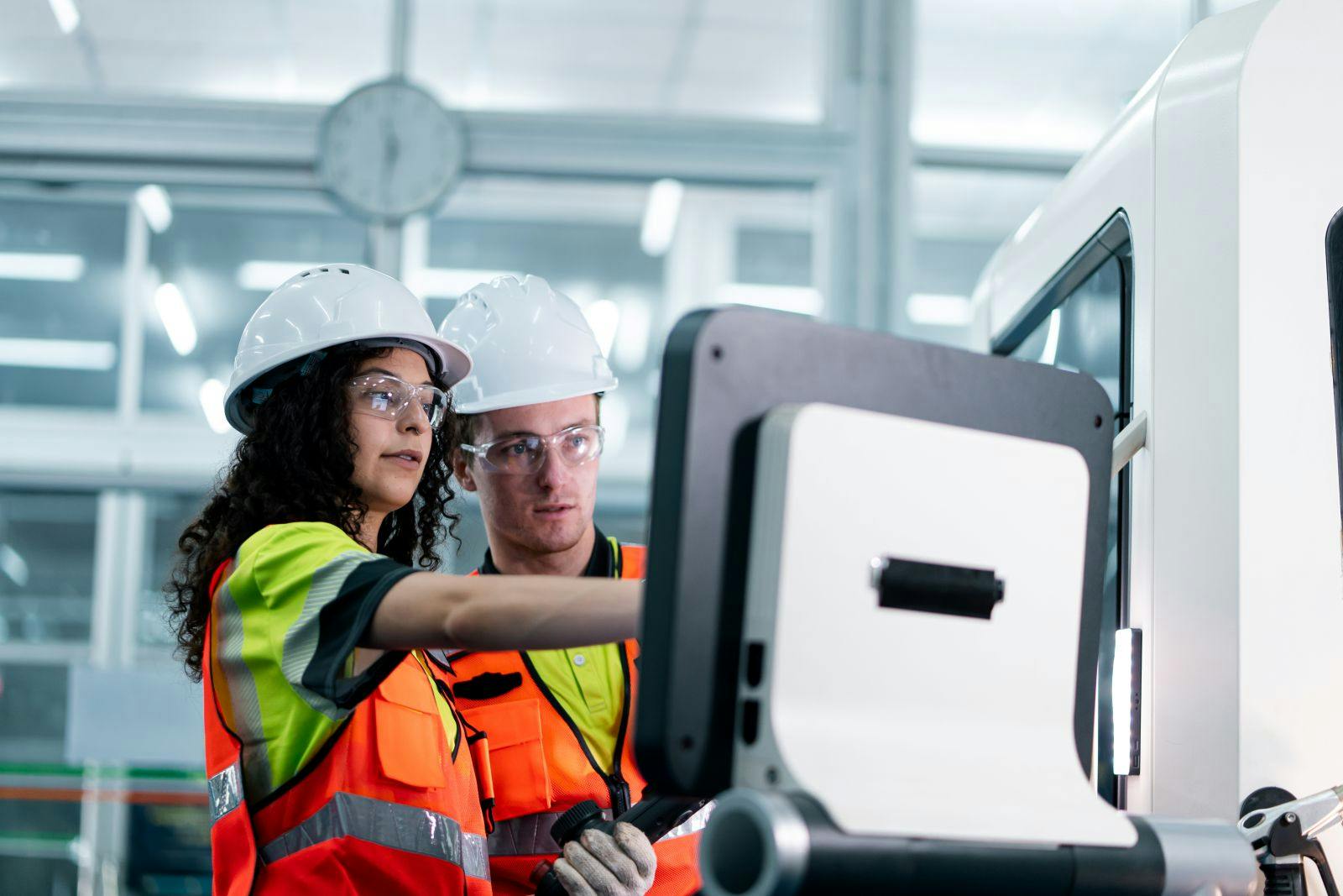 Two workers in orange vests are working on a machine. One of them is pointing at a screen