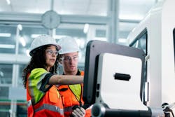 Two workers in orange vests are working on a machine. One of them is pointing at a screen Two workers in orange vests are working on a machine. One of them is pointing at a screen
