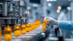 A gloved hand pours pills into a bottle on a conveyor belt in a pharmaceutical manufacturing facility. Multiple filled bottles are in a row.