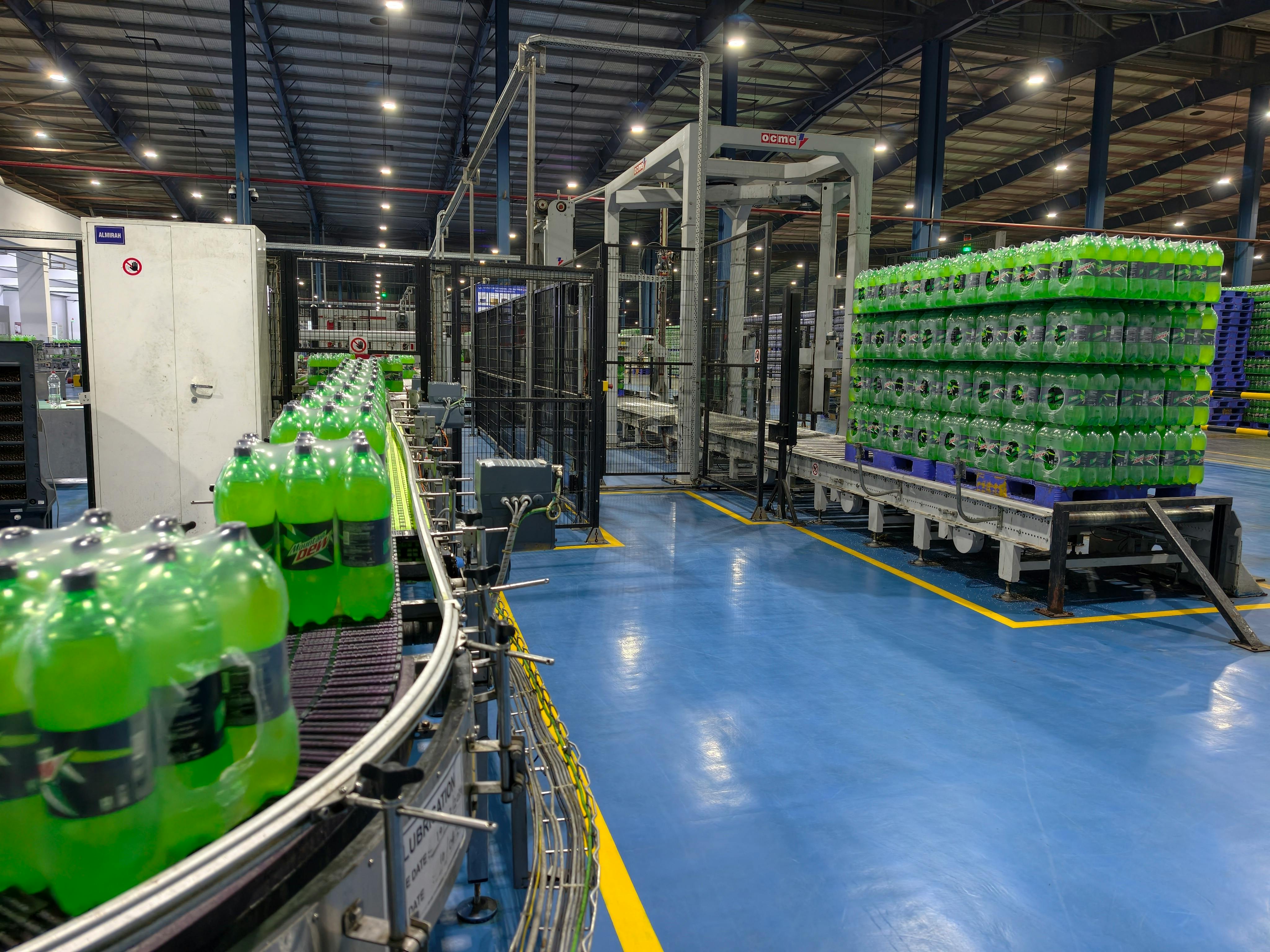 A modern beverage production line inside a large industrial factory, showcasing rows of green carbonated soft drink bottles moving along an automated conveyor system. The image highlights advanced manufacturing technology, with neatly stacked pallets of bottled drinks ready for distribution in the background. Bright overhead lighting illuminates the clean, organized facility, emphasizing efficiency, hygiene, and high-volume production.