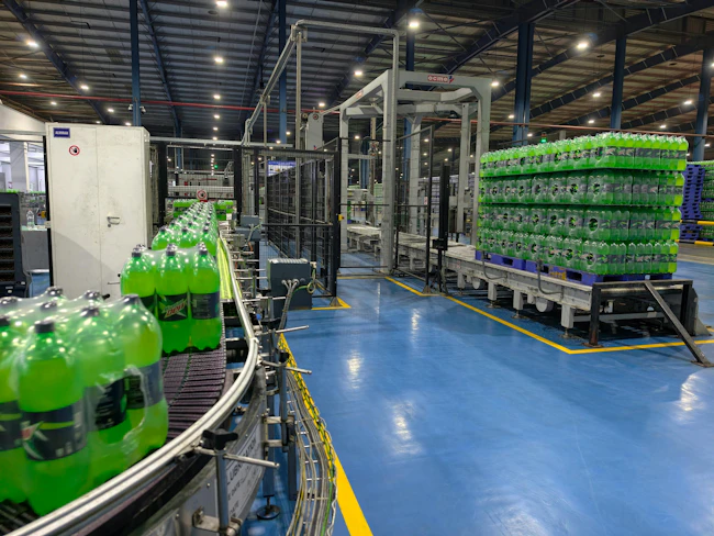 A modern beverage production line inside a large industrial factory, showcasing rows of green carbonated soft drink bottles moving along an automated conveyor system. The image highlights advanced manufacturing technology, with neatly stacked pallets of bottled drinks ready for distribution in the background. Bright overhead lighting illuminates the clean, organized facility, emphasizing efficiency, hygiene, and high-volume production.