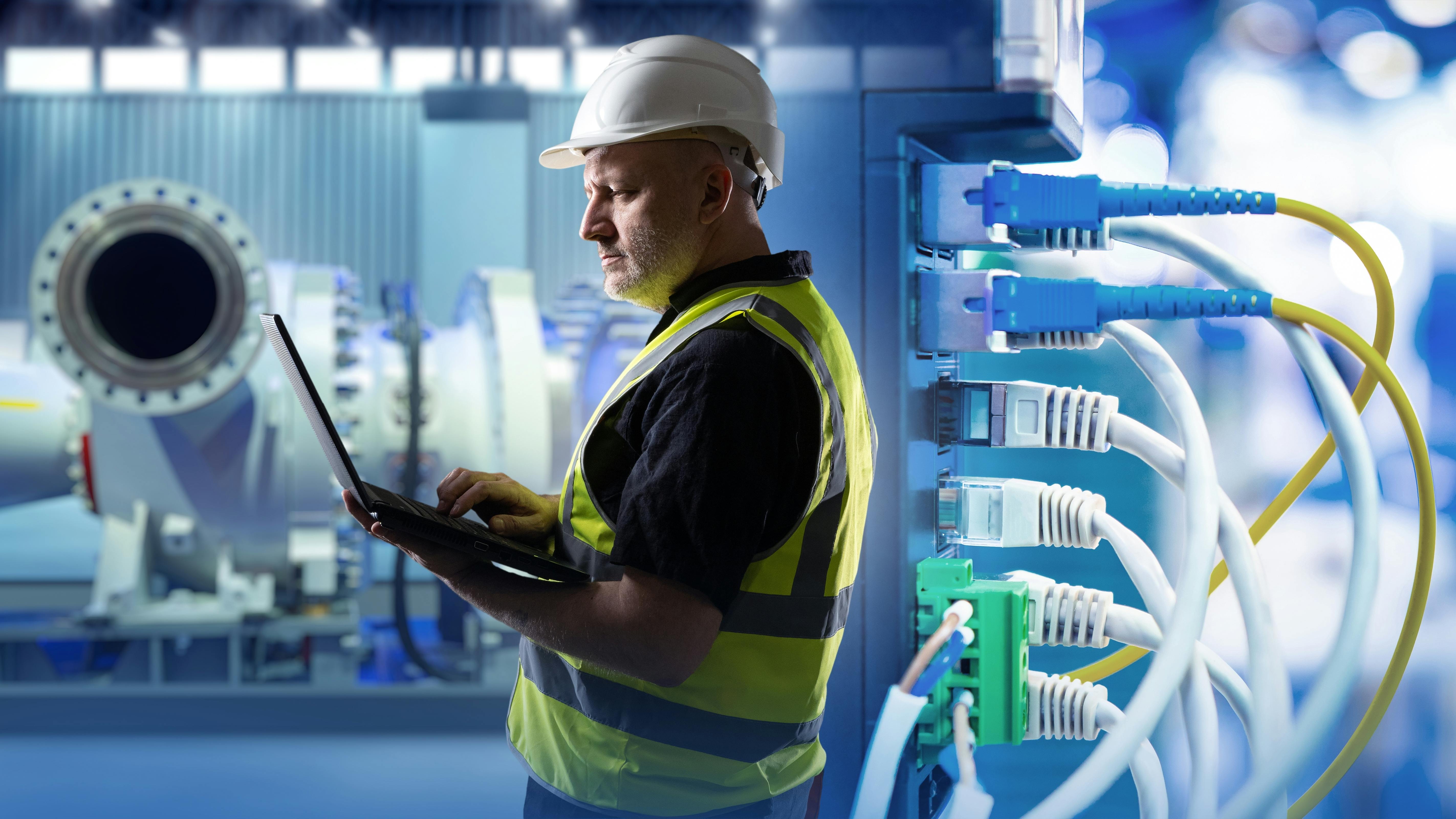 Industrial networking, equipment maintenance. Engineer uses laptop near network cables and machinery, monitoring systems and ensuring connectivity in production facility.