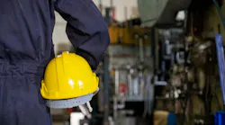 Back view close up of mechanic or worker holding yellow hard hat and wearing blue bear suit in factory with factory background.
