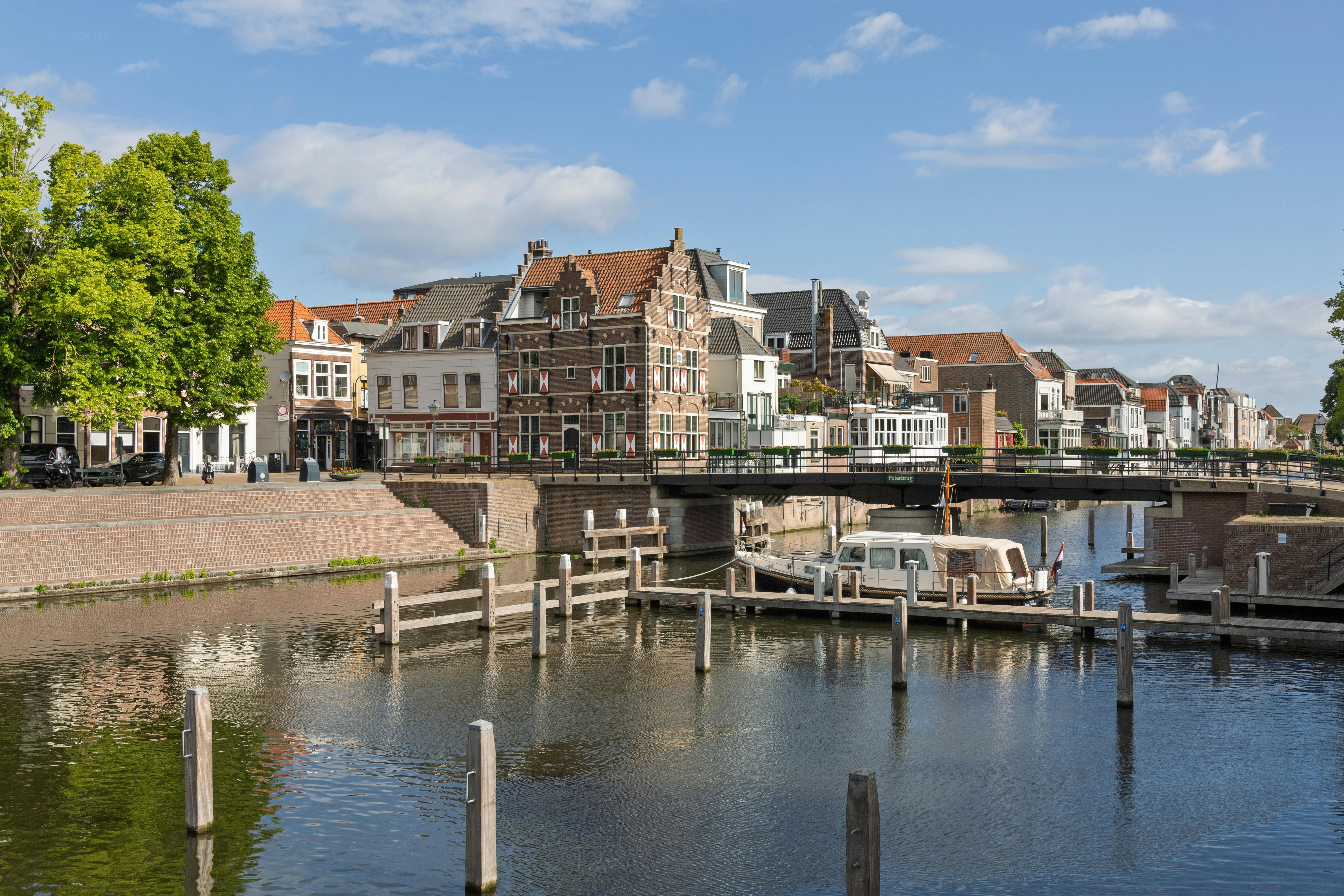 A picturesque scene in Gorinchem, Netherlands, showcasing traditional Dutch canal houses, the Peterbrug (Peter Bridge), and a house boat on the tranquil canal under a bright sky.