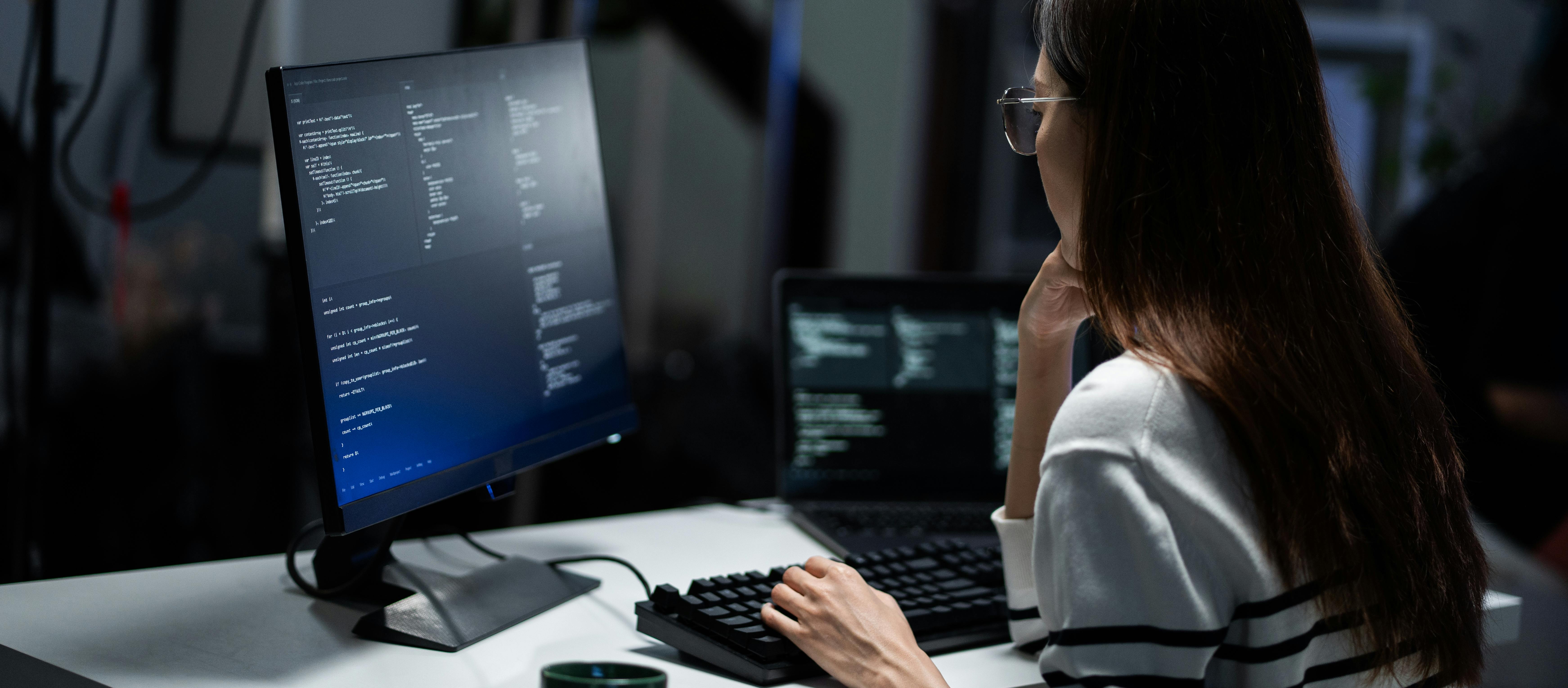 Rear view of a young programmer intensely reviewing code displayed on a monitor in a dark office.