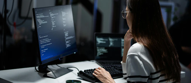 Rear view of a young programmer intensely reviewing code displayed on a monitor in a dark office.
