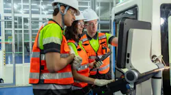 Three people wearing safety gear are standing in front of a computer monitor. One of the men is pointing at the screen