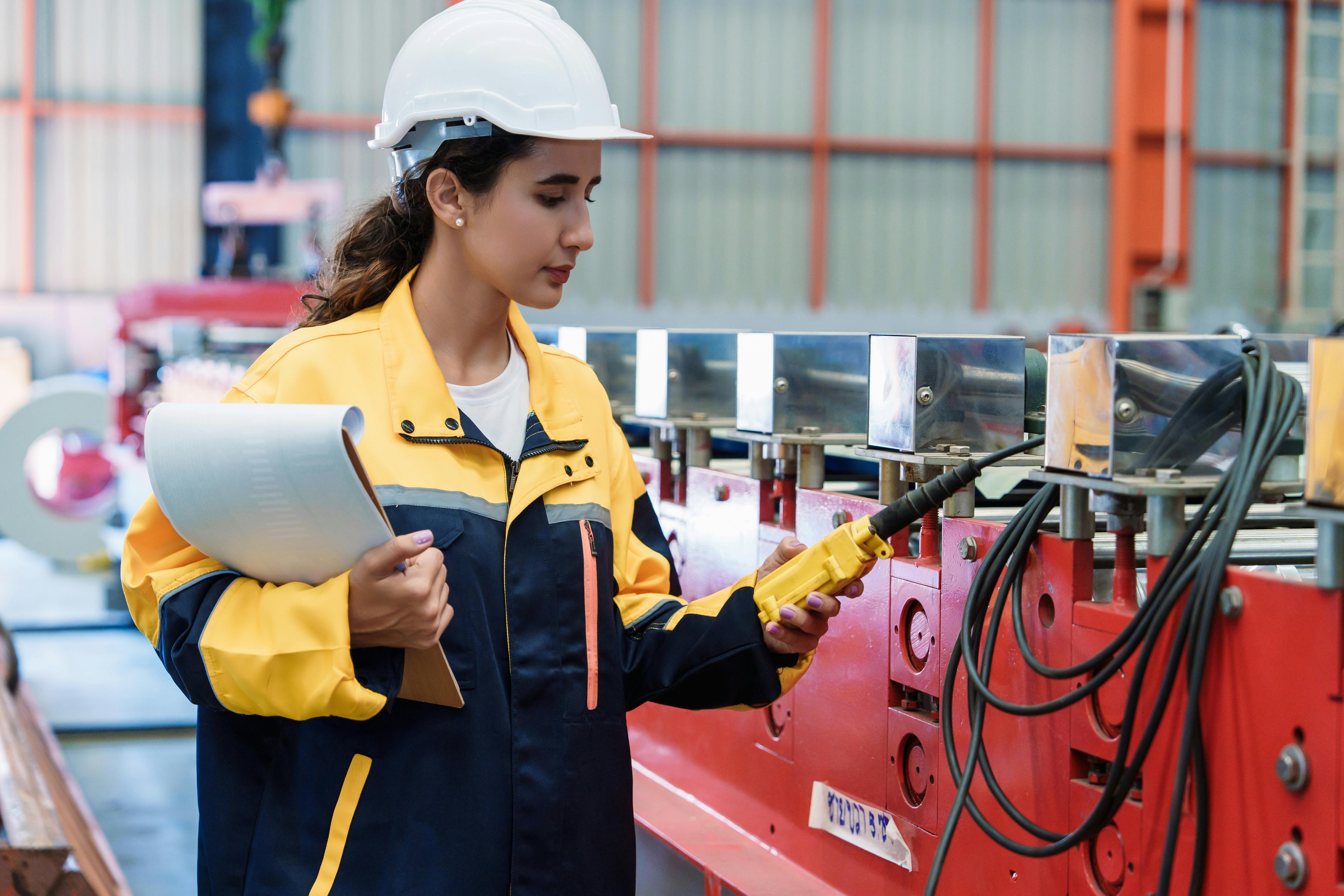 Female engineer carefully inspects roofing sheet production machine while holding blueprint at steel factory. Represents responsibility, accuracy, and industrial professionalism.