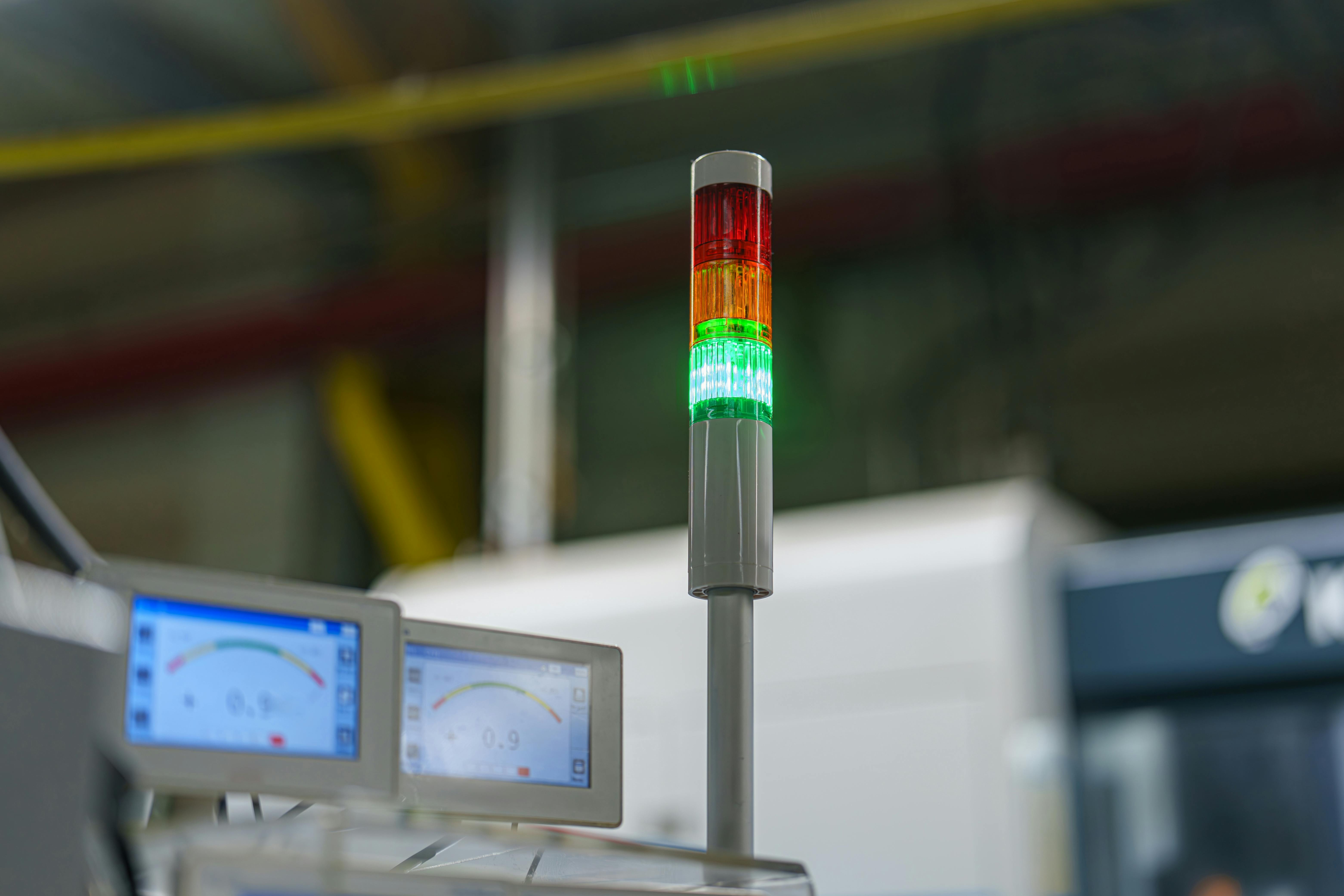 A signal tower shows a combination of red, yellow, and green lights in an industrial area. Screens displaying data are positioned near machinery. The setting is active with ongoing operations.