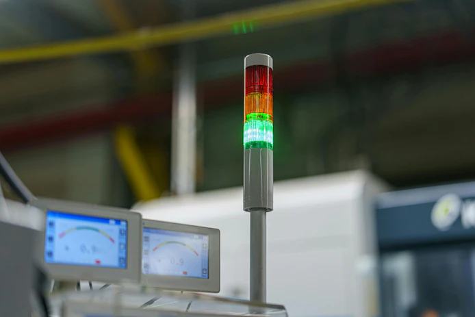 A signal tower shows a combination of red, yellow, and green lights in an industrial area. Screens displaying data are positioned near machinery. The setting is active with ongoing operations.