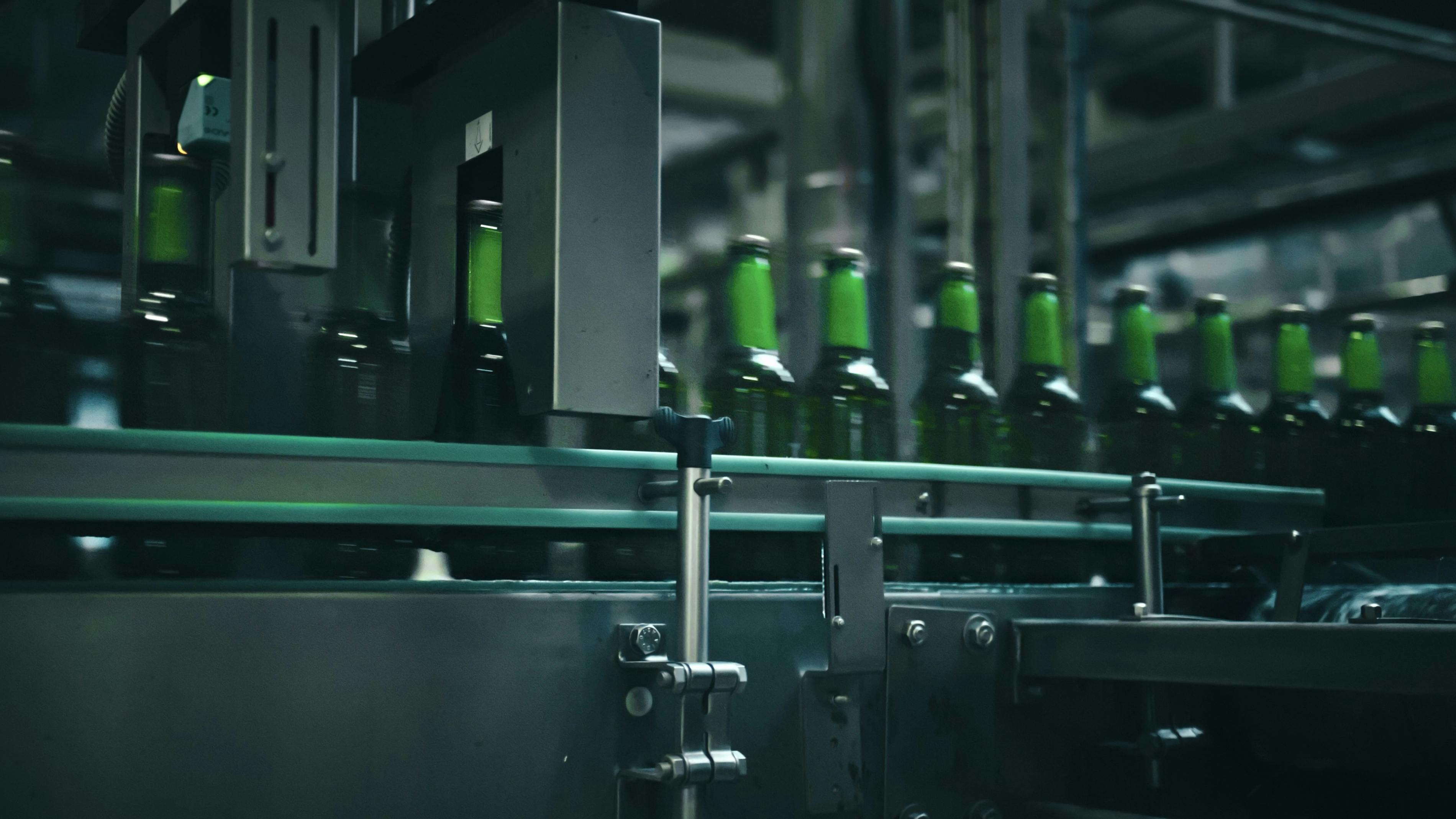 Rows of green glass bottles gliding along a conveyor belt inside an industrial brewery, moving through quality control machinery designed for efficient production and inspection
